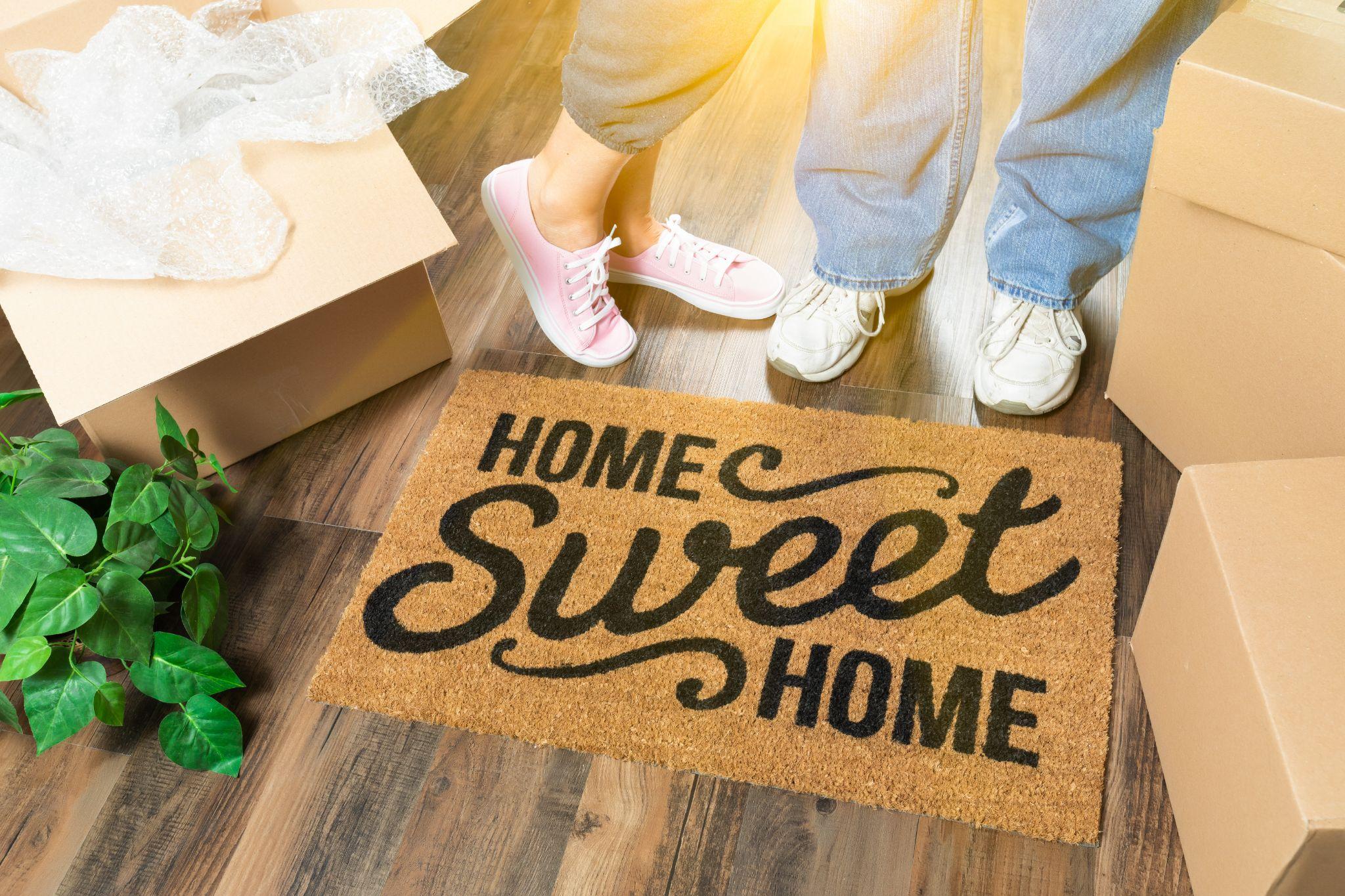 Home sweet home welcome mat with moving boxes, bubble wrap with a couple's feet 