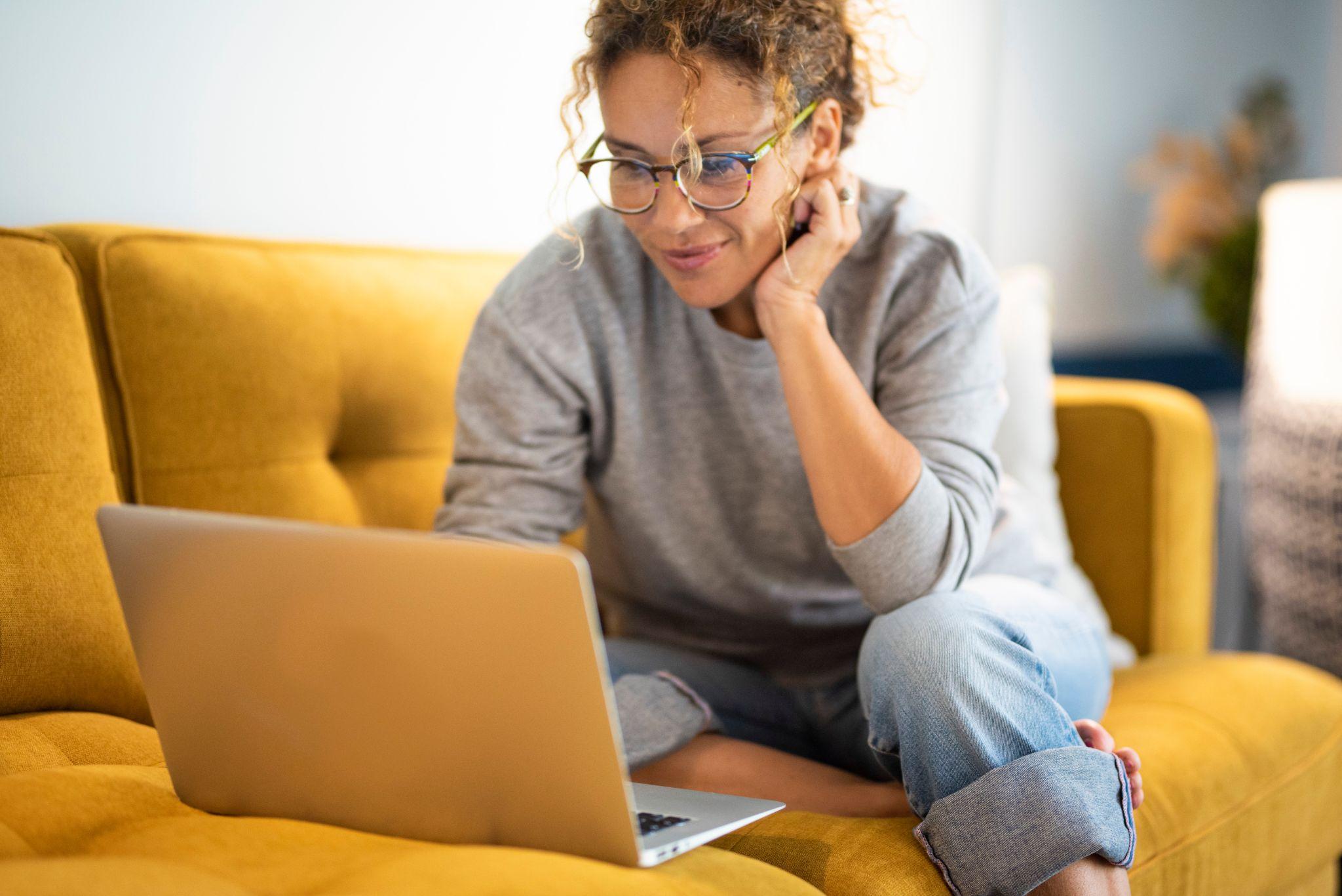 Female sitting on a yellow couch using laptop and internet connection and smile. 