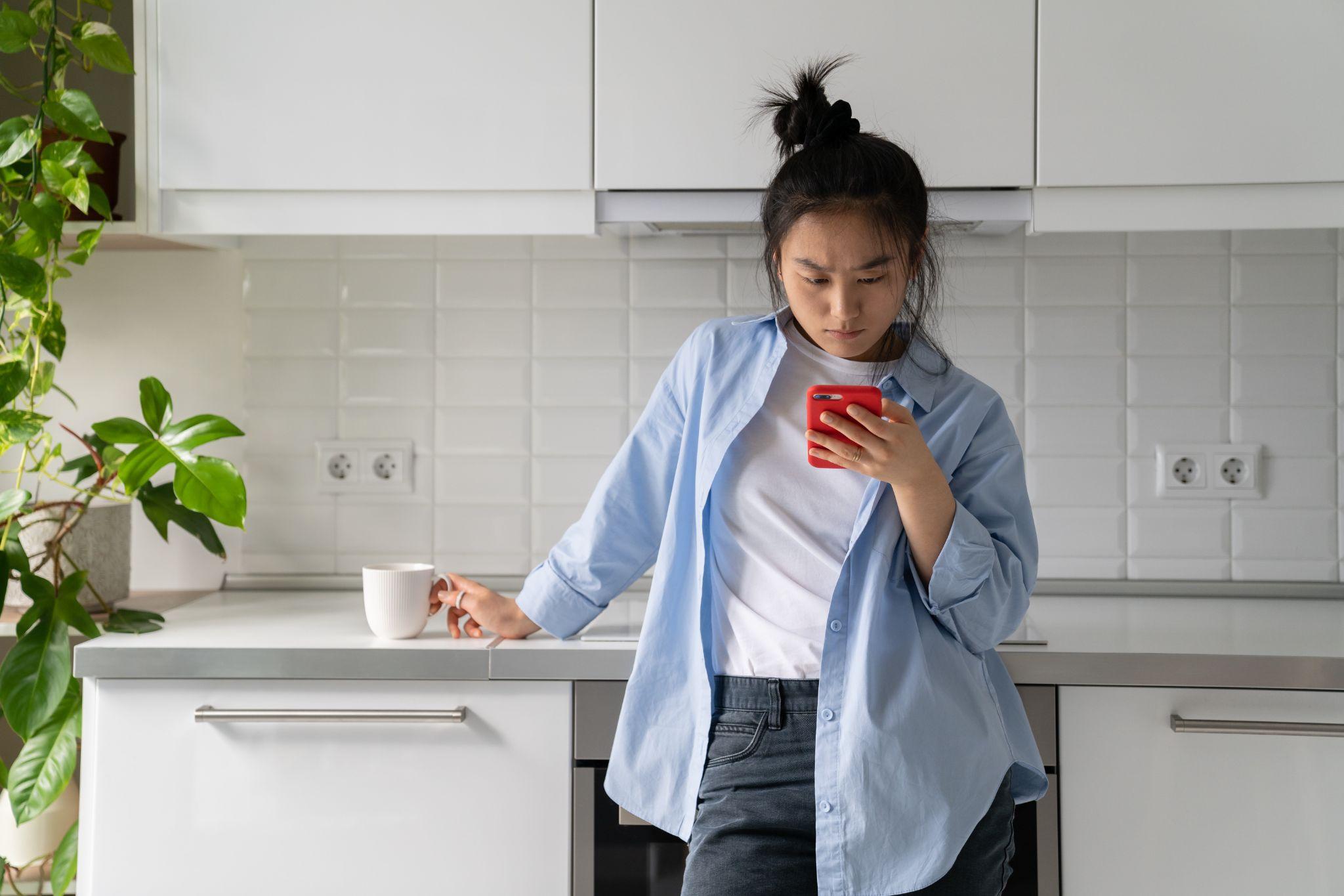 Upset young Asian woman standing in kitchen holding mobile phone